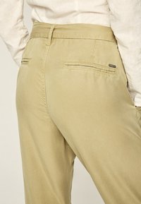 Pantaloni beige con una superficie liscia, dotati di due tasche posteriori e dettagli di cucitura discreti. La vita è piatta e presenta un passante per la cintura.