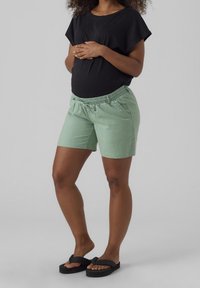 Personne enceinte portant un haut noir, un short vert clair et des tongs noires, debout avec les mains posées sur le ventre.