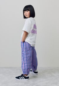 Witte T-shirt met paarse tekst, losse broek met roze en blauwe ruitjes en zwarte sneakers met witte strepen. Effen achtergrond.