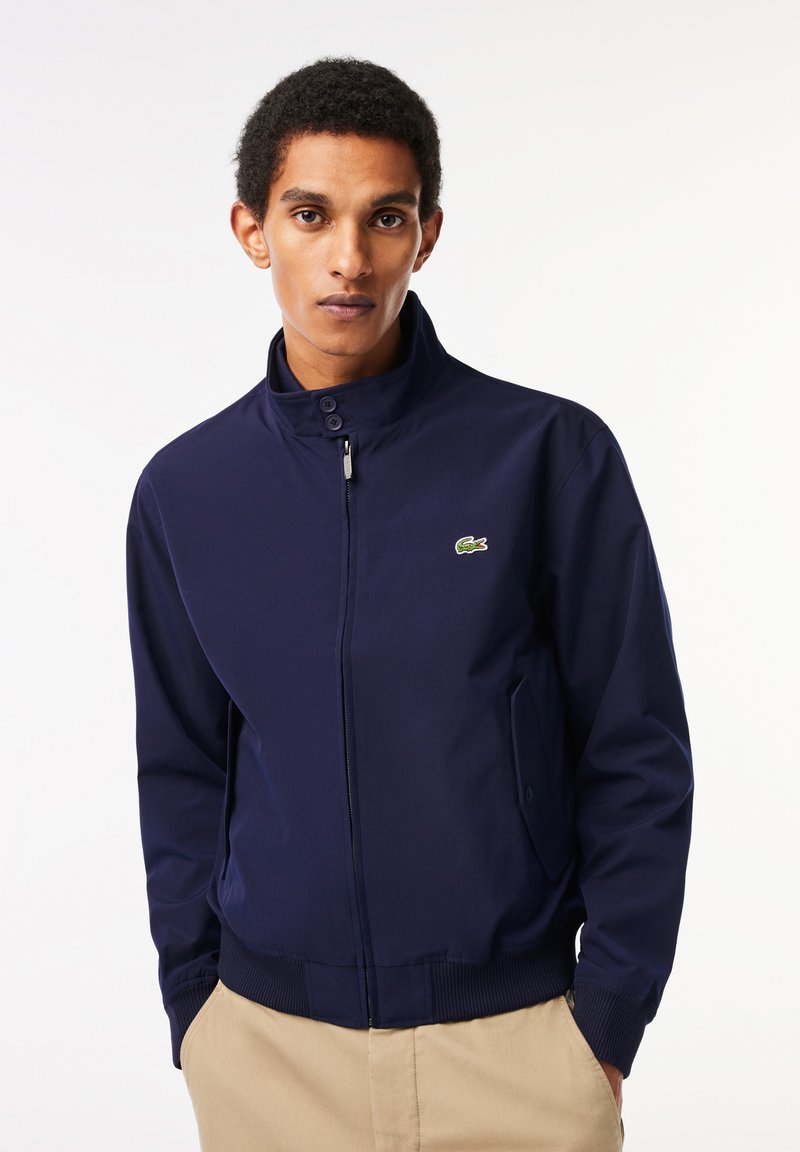Veste bomber bleu marine en tissu lisse avec un col montant, fermeture éclair à l'avant, poignets côtelés et un petit logo Lacoste sur la poitrine.