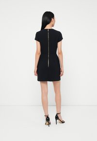 Mujer con cabello largo y negro, vestida con un vestido corto negro con un visible cremallera dorada que recorre la parte trasera y sandalias de tacón alto negras.