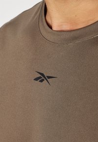 Camiseta marrón de manga corta hecha de una tela suave, con un logo negro en el pecho y un cuello redondo acanalado.