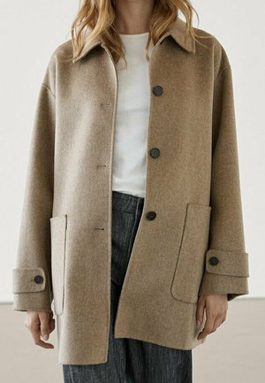 Donna che indossa un cappotto di lana beige con grandi tasche anteriori, bottoni neri e cinturini ai polsini sopra una camicia bianca e pantaloni grigi.