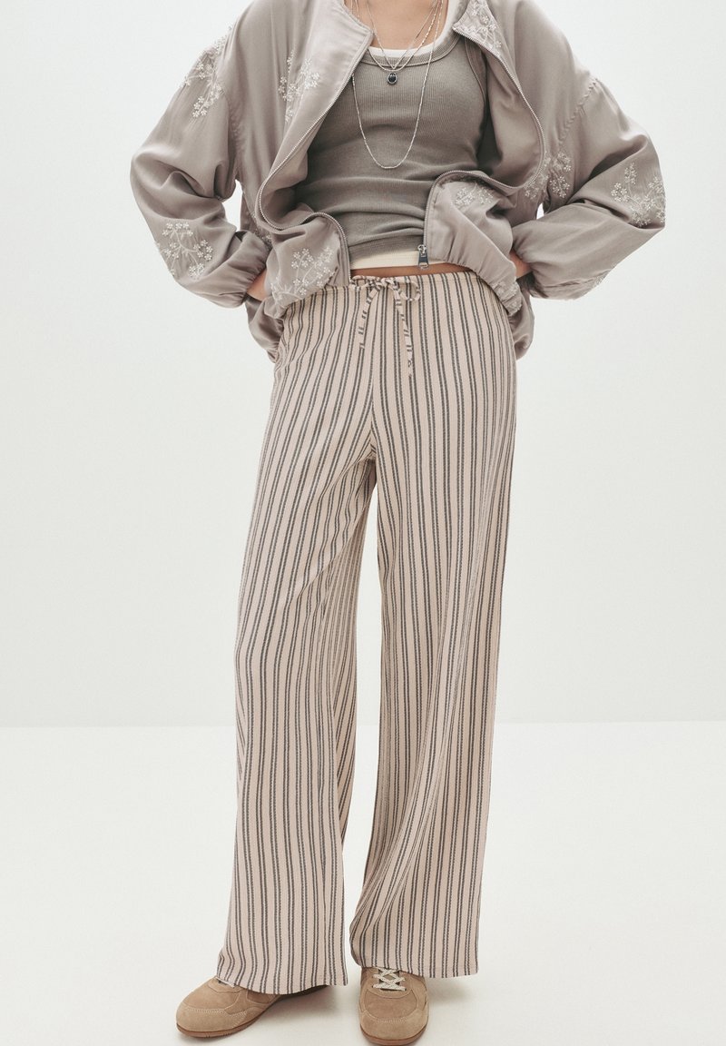 Personne portant un pantalon ample rayé beige et marron, un débardeur taupe, une veste brodée florale marron clair et des baskets beiges.