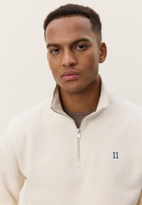 Μπεζ φλις μπλούζα με τεταρτημόριο φερμουάρ, ασημένιο φερμουάρ και πράσινο κεντημένο λογότυπο "II" στο στήθος. Ομαλή υφή και άνετη εφαρμογή.