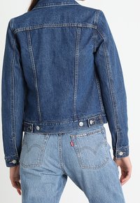 Person wears dark blue denim jacket and light blue jeans, shown from the back, standing against a plain white background.