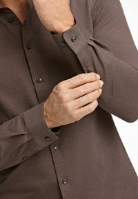 Camisa de botones de manga larga color marrón con una textura suave. Presenta botones oscuros, puños ajustables y un diseño de cuello clásico.