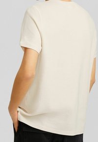 Beige t-shirt med kort ärm i strukturerad bomull, med rund halsringning och avslappnad passform med sidsömmar, bärs med händerna i fickorna.