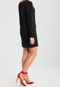 Femme portant une robe mini noire à manches longues avec des détails de manches volantes et des chaussures à talons aiguilles rouges à bride de cheville, se tenant de côté.