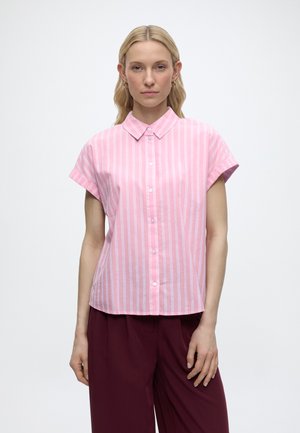 Femme portant une chemise à manches courtes rose clair à boutons avec des rayures blanches verticales et un pantalon rouge foncé, debout devant un fond uni.