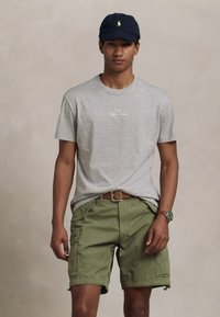 Jovem vestido com uma t-shirt cinza de mangas curtas, calções cargo verde oliva com cinto, boné de basebol azul-escuro e relógio de pulso, posando contra um fundo neutro.