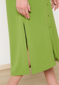 Abito midi verde realizzato in tessuto morbido con uno spacco laterale e dettagli di bottoni. Texture liscia, vestibilità comoda, design semplice.