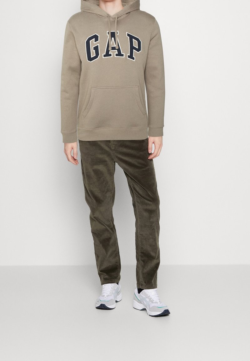 Felpa color khaki con logo "GAP" in blu navy, tasca frontale, abbinata a pantaloni in velluto a coste color oliva. Il modello indossa scarpe da ginnastica bianche e grigie.