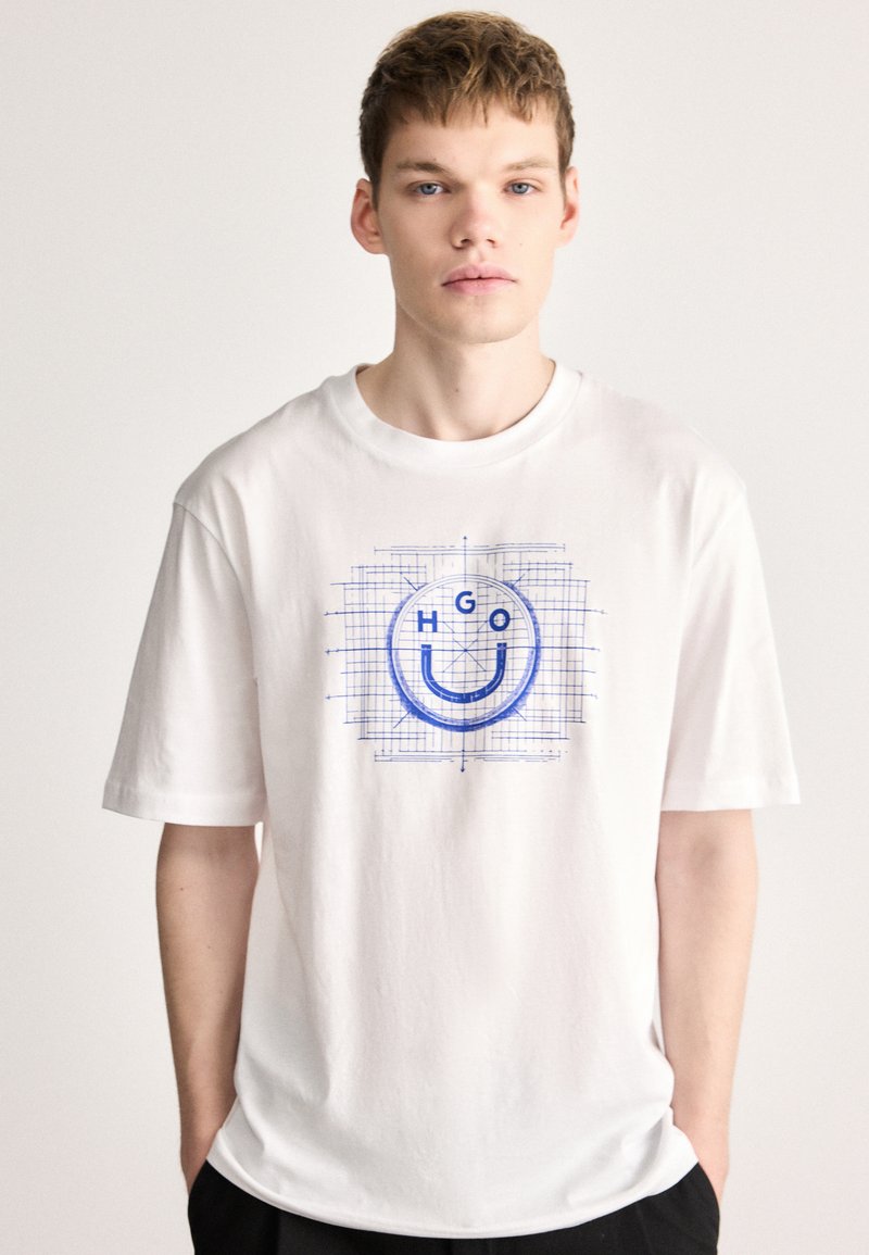 Camiseta blanca de algodón con un diseño gráfico azul que incluye una cara sonriente y las letras "HGO" sobre un fondo de cuadricula. Manga corta, corte relajado.