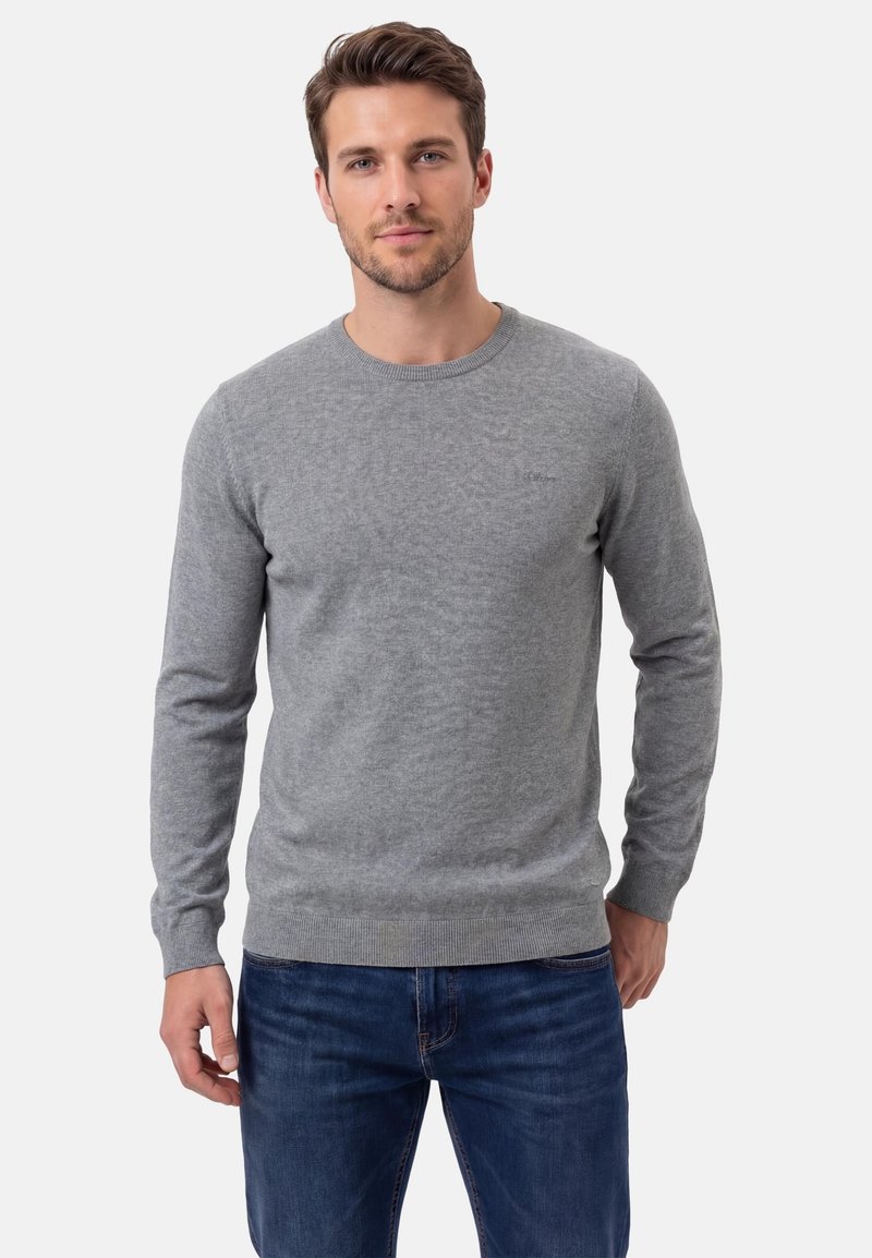 Mann trägt einen hellgrauen Langarmpullover und blaue Jeans, steht vor einem schlichten weißen Hintergrund.