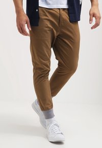 Pantaloni marroni svasati con polsini elastici, realizzati in un materiale morbido, abbinati a sneakers bianche e calzini grigi. Dettagli sottili delle cuciture visibili.