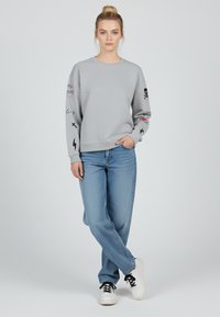Felpa grigia con stampe grafiche sulle maniche, scollo a girocollo, abbinata a jeans a gamba dritta blu chiaro e sneaker bianche con lacci neri.