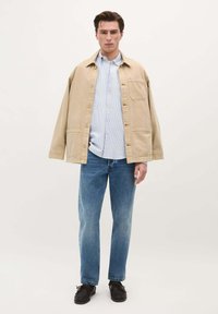 Chaqueta de mezclilla oversize beige sobre una camisa de rayas azul claro, combinada con jeans azul claro y zapatos negros. Tejido texturizado y diseño informal.