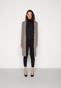 Long, light brown cardigan with button front, worn over a black turtleneck, paired with black skinny pants and black heels. Smooth texture.