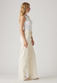 Calças de ganga creme com corte amplo, cintura alta e bolsos frontais. O tecido tem uma textura suave com detalhes de costuras contrastantes.