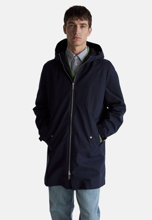 Joven vestido con un abrigo con capucha azul marino oscuro sobre un suéter verde y una camisa a rayas, con las manos en los bolsillos del abrigo, de pie frente a un fondo blanco.