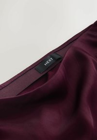 Donker bordeaux satijnen stof met een gladde textuur, voorzien van een zwart label met "NEXT" dat in de halslijn is genaaid.