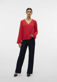 Blusa rossa a maniche lunghe con scollo a V e chiusura a bottoni, caratterizzata da sottili punti testurizzati. Abbinata a jeans a gamba larga blu scuro e scarpe nere.