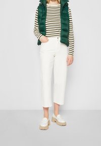 Zielona kamizelka puchowa na cienkich paskach, zestawiona z białymi spodniami 3/4 i masywnymi beżowymi mokasynami. Gładkie tekstury i kontrastowe kolory.