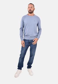 Uomo con barba corta che indossa un maglione blu chiaro, jeans blu e sneakers bianche, in piedi contro uno sfondo neutro.