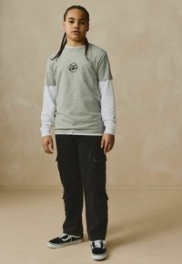 T-shirt en coton gris sur une chemise à manches longues blanche, associée à un pantalon cargo noir et des baskets noires, avec de multiples poches et un logo.