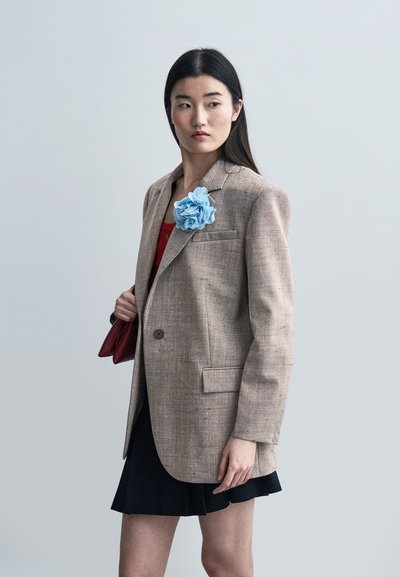 Femme portant un blazer beige avec une épingle fleur bleue, un haut rouge, une jupe noire et un sac à main bordeaux, regardant vers sa gauche sur un fond gris uni.