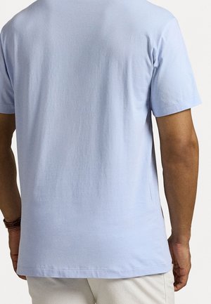 Homme portant un t-shirt bleu clair à manches courtes et un short blanc, vu de dos sur un fond uni.