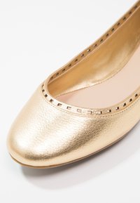 Gros plan d'une chaussure plate de ballet dorée métallique avec des découpes carrées le long du bord et un bout arrondi.