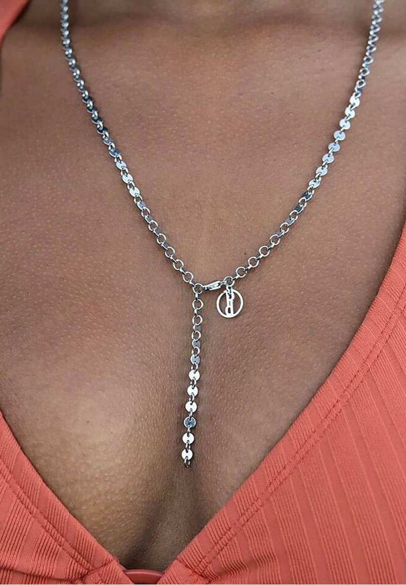 Collier chaîne en argent avec un petit pendentif circulaire, porté sur une peau brune et un haut côtelé à col en V orange.