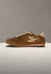 Καφέ σουέντ αθλητικό παπούτσι με λευκά κορδόνια, λευκή λεπτομέρεια στη φτέρνα, κόκκινη ρίγα κοντά στη σόλα και καλλιγραφικό λογότυπο "ZV" στο πλάι.