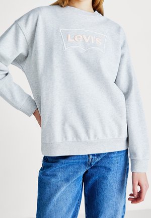 Personne portant un sweat-shirt Levi's gris clair et un jean bleu, main sur la hanche, debout devant un fond clair uni.