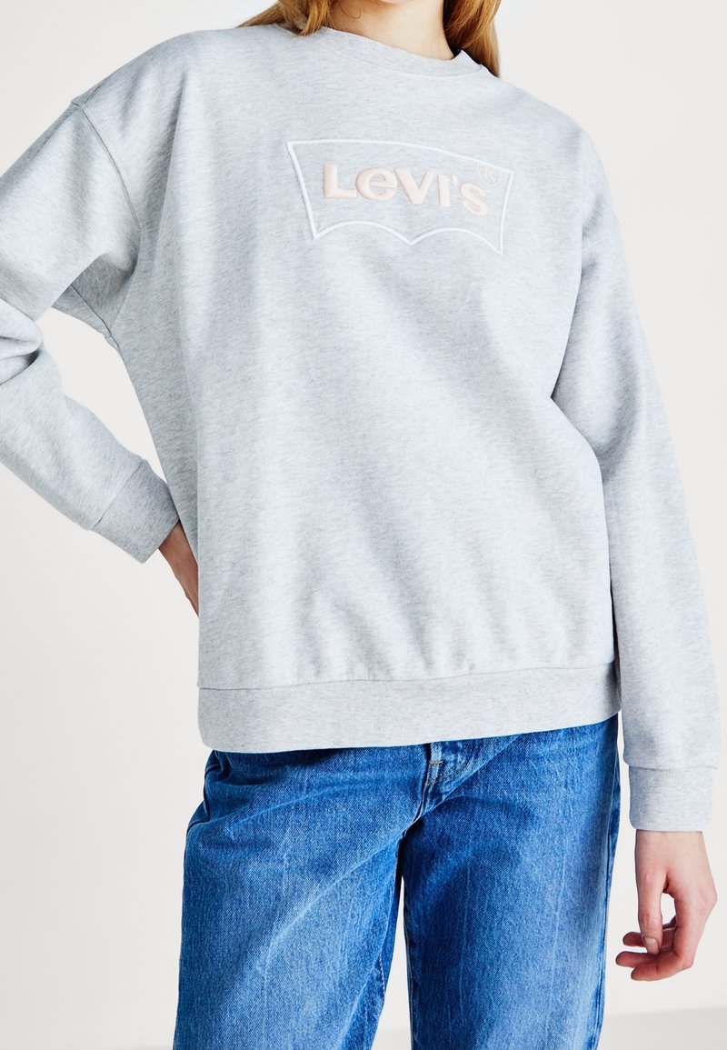 Personne portant un sweat-shirt Levi's gris clair et un jean bleu, main sur la hanche, debout devant un fond clair uni.