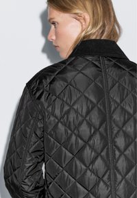 Black quilted jacket with a textured, shiny finish, featuring a ribbed collar and diamond-patterned stitching. Back view displayed.