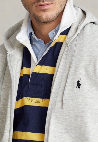 Grå dragkedjehoodie över en marinblå och gulrandig pikétröja. Detaljer inkluderar krage, ficka och broderad logotyp på hoodien.