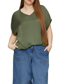 Oliwkowa bluzka z krótkim rękawem i dekoltem w kształcie litery V. Połączona z szerokimi niebieskimi dżinsami z przednimi kieszeniami. Gładka struktura.