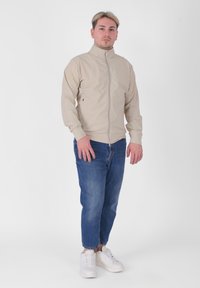 Giacca leggera beige con chiusura a zip, colletto alto e polsini elasticizzati, abbinata a jeans slim-fit blu scuro e sneakers bianche.