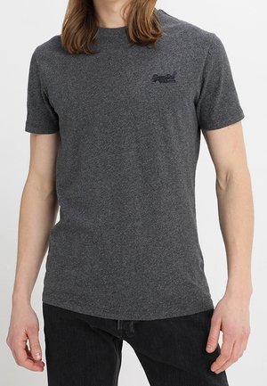 Mężczyzna w dopasowanej, ciemnoszarej koszulce z krótkim rękawem z małym czarnym logo po lewej stronie klatki piersiowej oraz w czarnych spodniach na białym tle.