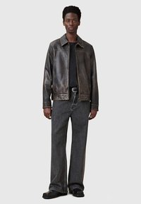 Giacca di pelle marrone consumata con scollo a collo, chiusura a zip e maniche lunghe, abbinata a jeans neri a gamba larga e calzature nere.