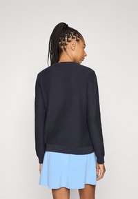 Tengerészkék kötött pulóver kerek nyakkal és bordázott mandzsettákkal, világoskék, bővülő szoknyával párosítva, texturált anyagot mutatva.