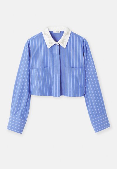 Camisa azul a rayas cortada con mangas largas, que presenta un color blanco bordado en el cuello y dos bolsillos frontales. Textura de tela suave.