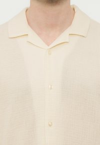 Κοντινό πλάνο ενός ατόμου που φοράει ένα ανοιχτό κρεμ πουκάμισο με υφή, κουμπωτό, με γιακά καμπς, δείχνοντας το λαιμό και το πάνω μέρος του στήθους.