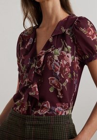 Blommig blus i djup burgundy med volangkrage och korta puffärmar. Har ett flerfärgat blommönster på lättviktig tyg.