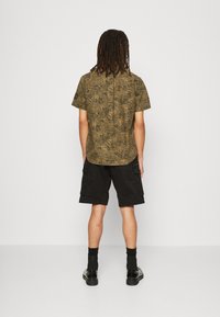 Korte mouwen, olijfgroene blouse met palmbladpatroon, gecombineerd met zwarte cargo shorts. De stof lijkt lichtgewicht met een relaxte pasvorm.