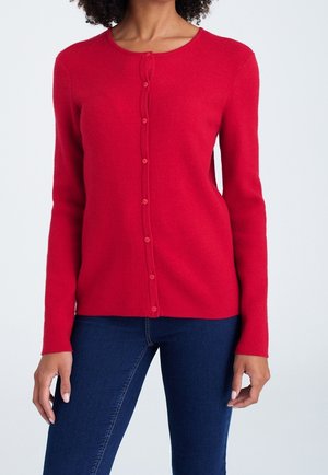 Vrouw die een nauwsluitend, felrood vest met knopen draagt, met lange mouwen en donkerblauwe strakke jeans, tegen een effen achtergrond.