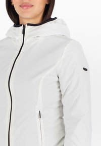 Witte capuchonjack met een gladde, glanzende textuur. Heeft een zwarte rits en accenten. Bevat een ritszak aan de linkerkant.